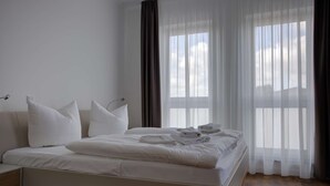 Cama bem arrumada com lençóis e almofadas brancos, mesa de cabeceira com abajur e cortinas leves que deixam entrar luz natural.