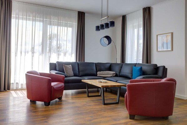 A modern living room with a dark leather sofa, two red armchairs, a wooden coffee table, and a floor lamp. - Villa Zeitlos - Vacation Apartment 11 (Börgerende-Rethwisch)