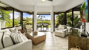Un vaste espace de vie comprenant un grand canapé, une table basse et un téléviseur. À l’extérieur de la fenêtre, on trouve une table à manger avec des chaises, et la pièce est dotée de grandes fenêtres offrant une vue sur la verdure.