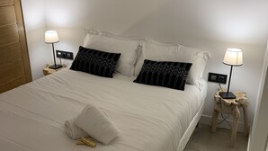 A neatly made bed with white linens, two black patterned pillows, and a folded towel on the bed. There are two bedside tables with lamps, one on each side of the bed. - Comfy Pinoche Proche Plage (Saint-Malo)