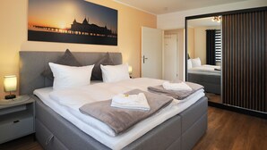 A modern hotel room with a large bed, a nightstand, a lamp, a painting of a sunset, a mirror, and a sliding door. - Fewo Sonnenblick - Fewo Sonnenblick (Heringsdorf)