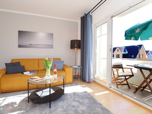 Ein modernes Wohnzimmer mit einer Couch, einem Glastisch, einer Lampe und Blick auf einen Balkon mit Sitzgelegenheit.