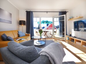 Un salon comprenant un canapé, des fauteuils, une table basse, un téléviseur et un balcon donnant sur des maisons.