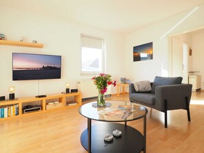 Ein modernes Wohnzimmer mit Flachbildfernseher, rundem Glastisch, grauem Sessel, Holzregal und Fenster mit Blick auf einen Steg.