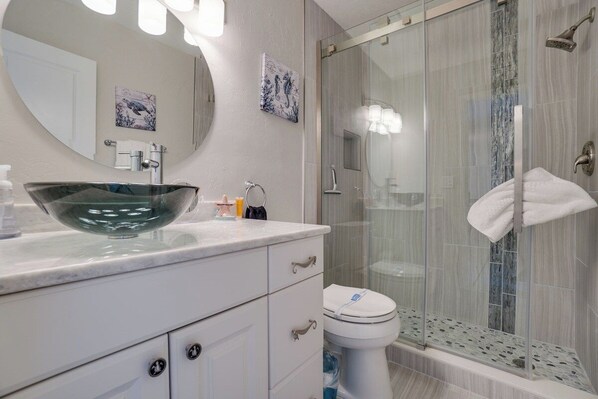 Un baño moderno con cabina de ducha de vidrio, inodoro blanco y un gran espejo ovalado sobre el lavamanos.