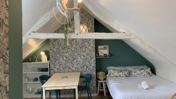 A cozy attic bedroom with a sloped ceiling, a wooden table, two chairs, a bed with floral pillows, a small stool, and a hanging light fixture. - Jungle Pinoche Proche Plage (Saint-Malo)
