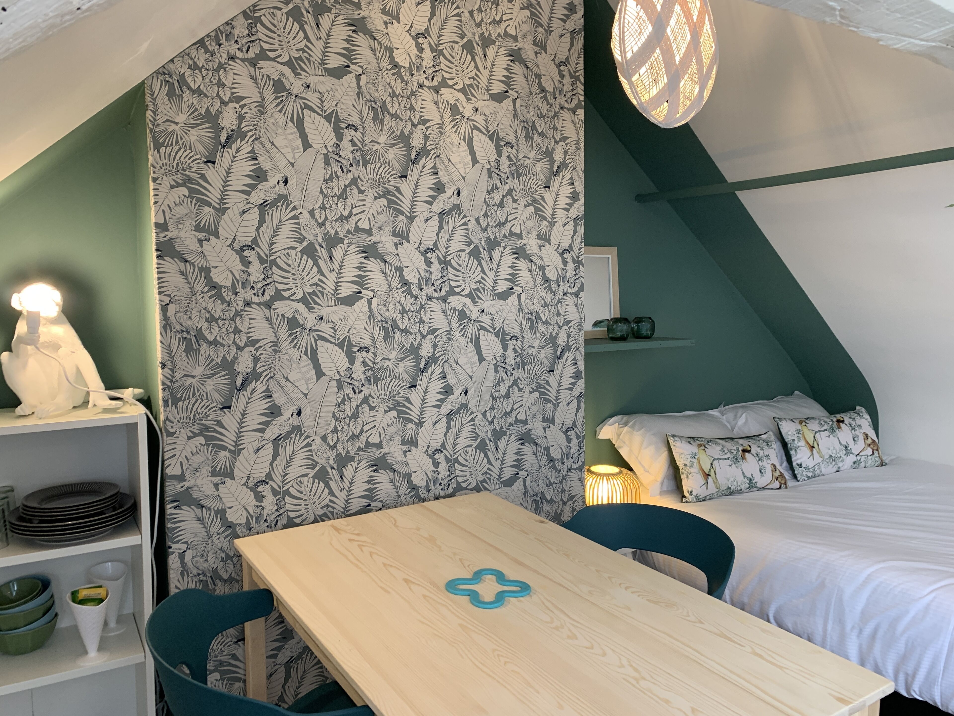 Una habitación acogedora con una mesa de madera, sillas color turquesa, una cama con almohadillas estampadas con flores y una pared con un diseño de hojas.