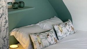 A bedroom with a bed, pillows, a nightstand, a lamp, and a shelf with decorative items. - Jungle Pinoche Proche Plage (Saint-Malo)