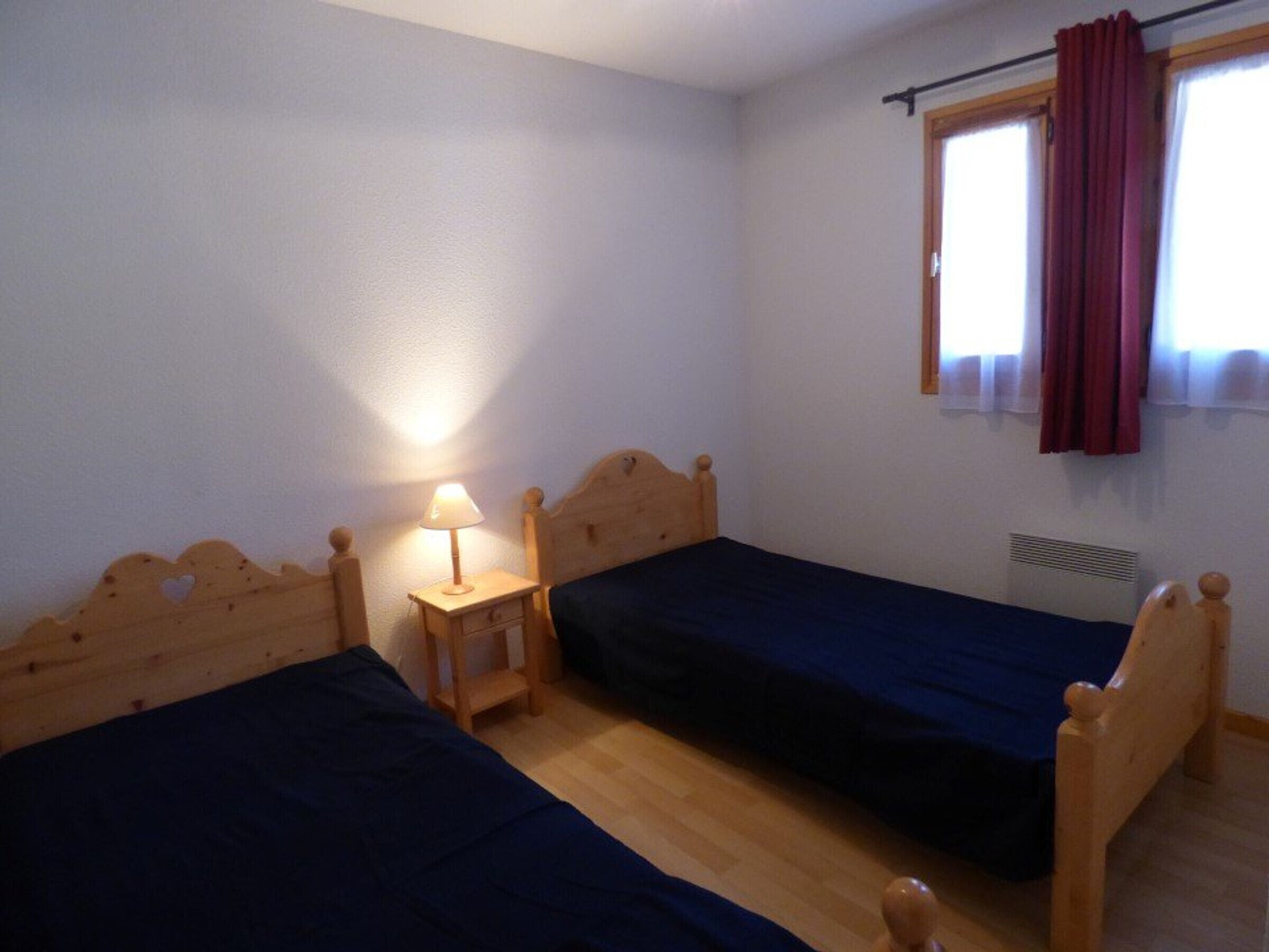 A small, simple room with two wooden beds, a nightstand with a lamp, and a window with curtains.