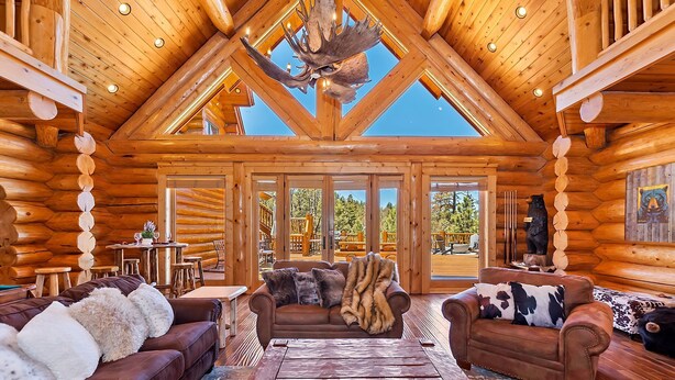 A log cabin interior with a high ceiling, wooden beams, a large chandelier, and a view of the outdoors.