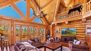 A log cabin interior with a large fireplace, leather sofas, a wooden coffee table, and a balcony with a bear statue.