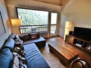 Una sala de estar moderna con sofá, una mesa de centro, televisor y un balcón con vistas al bosque.