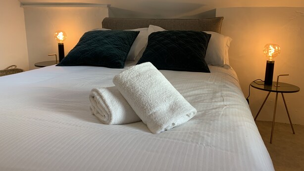 A neatly made bed with white linens, two dark pillows, and a rolled white towel. There are two bedside lamps with exposed bulbs on either side of the bed. - Simply Pinoche Room Proche Plage (Saint-Malo)