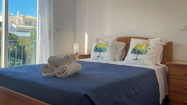 Una cama bien tendida con una colcha azul, dos almohadas con diseños de árboles y toallas enrolladas a un lado.
