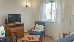 Una sala de estar con una televisión sobre un mueble de madera, un sofá, un sillón con cojín, una mesita con mandos a distancia y una ventana con cortinas.