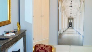 A hallway with a long corridor, a white wardrobe, a marble fireplace, and a red upholstered chair.