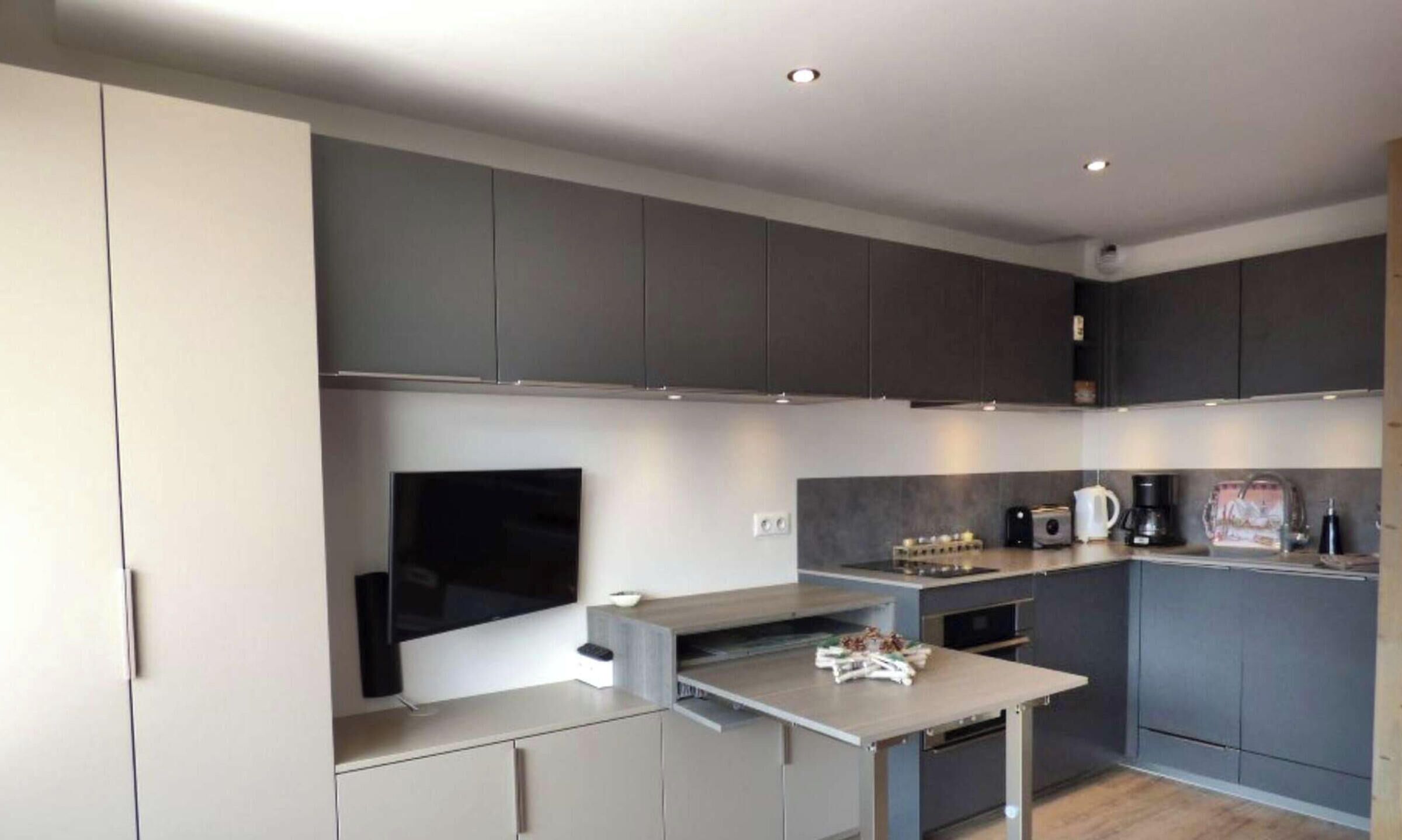 Una cocina moderna con gabinetes grises, horno empotrado, cafetera y televisor de pantalla plana.