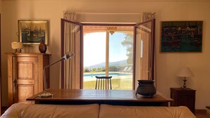 A room with a bed, a desk with a lamp, a view of a pool and mountains, and two paintings on the walls. - An oasis of peace and quite with sea view on Cannes bay (Peymeinade)