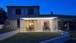 A modern house with a covered patio area featuring a dining table and chairs, a bench, and a small garden. - An oasis of peace and quite with sea view on Cannes bay (Peymeinade)