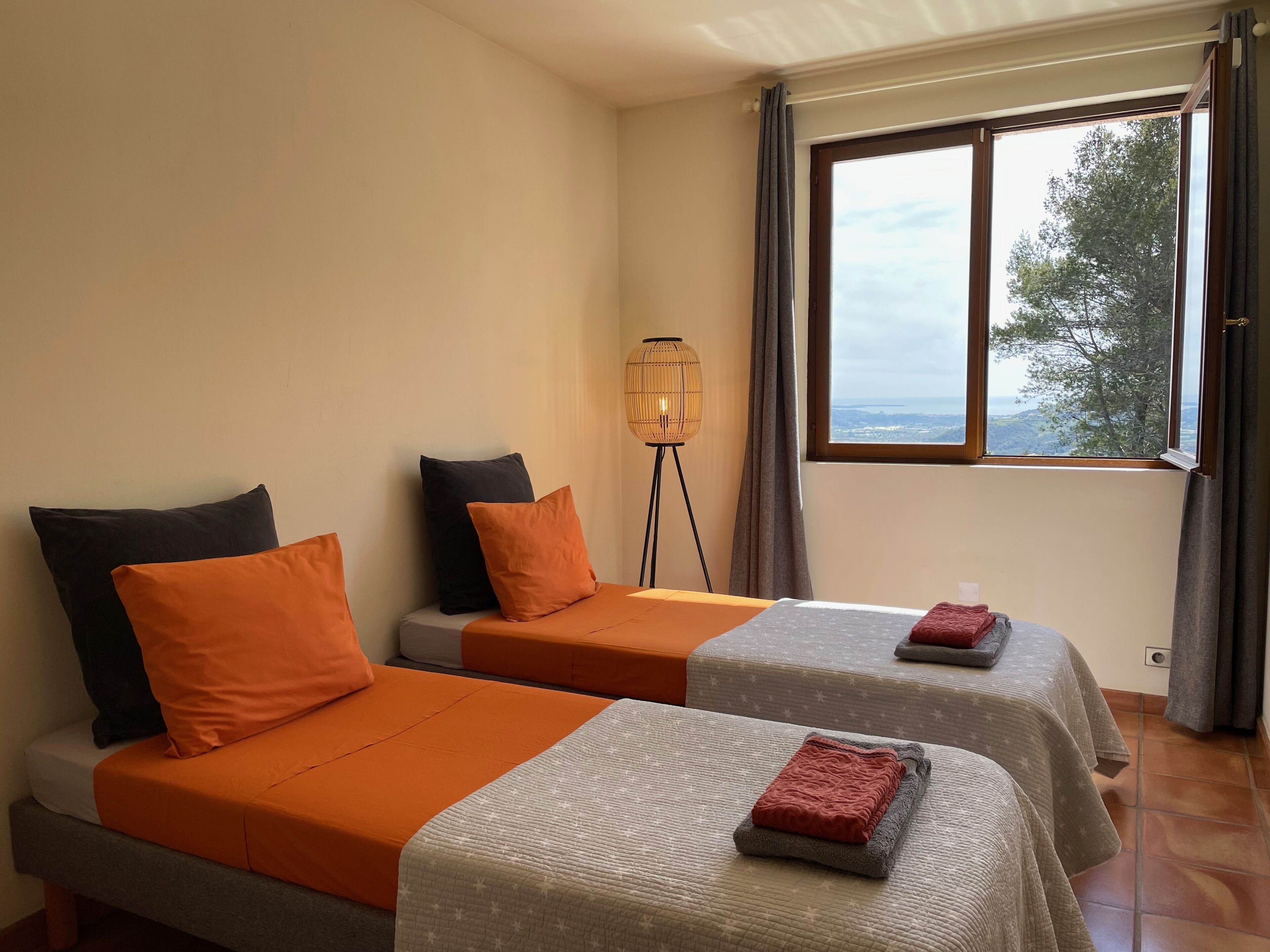 Quarto de hotel com duas camas, cada uma com colcha e almofadas laranja, uma luminária de cabeceira e uma janela com vista para árvores e colinas.