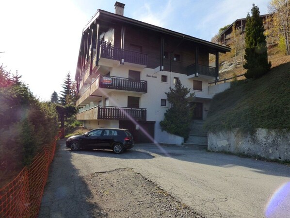 A multi-story building with balconies, a car parked in front, and a hilly background.