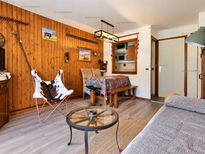 A cozy room with wooden walls, a dining table with a colorful tablecloth, a leather chair, a round glass table, and a bed.