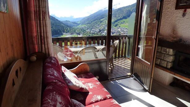 A balcony with a wooden bench and cushions, a glass door leading to a scenic view of a valley, and a fireplace.