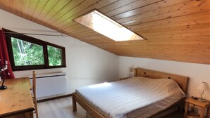 A bedroom with a wooden ceiling, a bed, a desk, and a window with a view of trees.