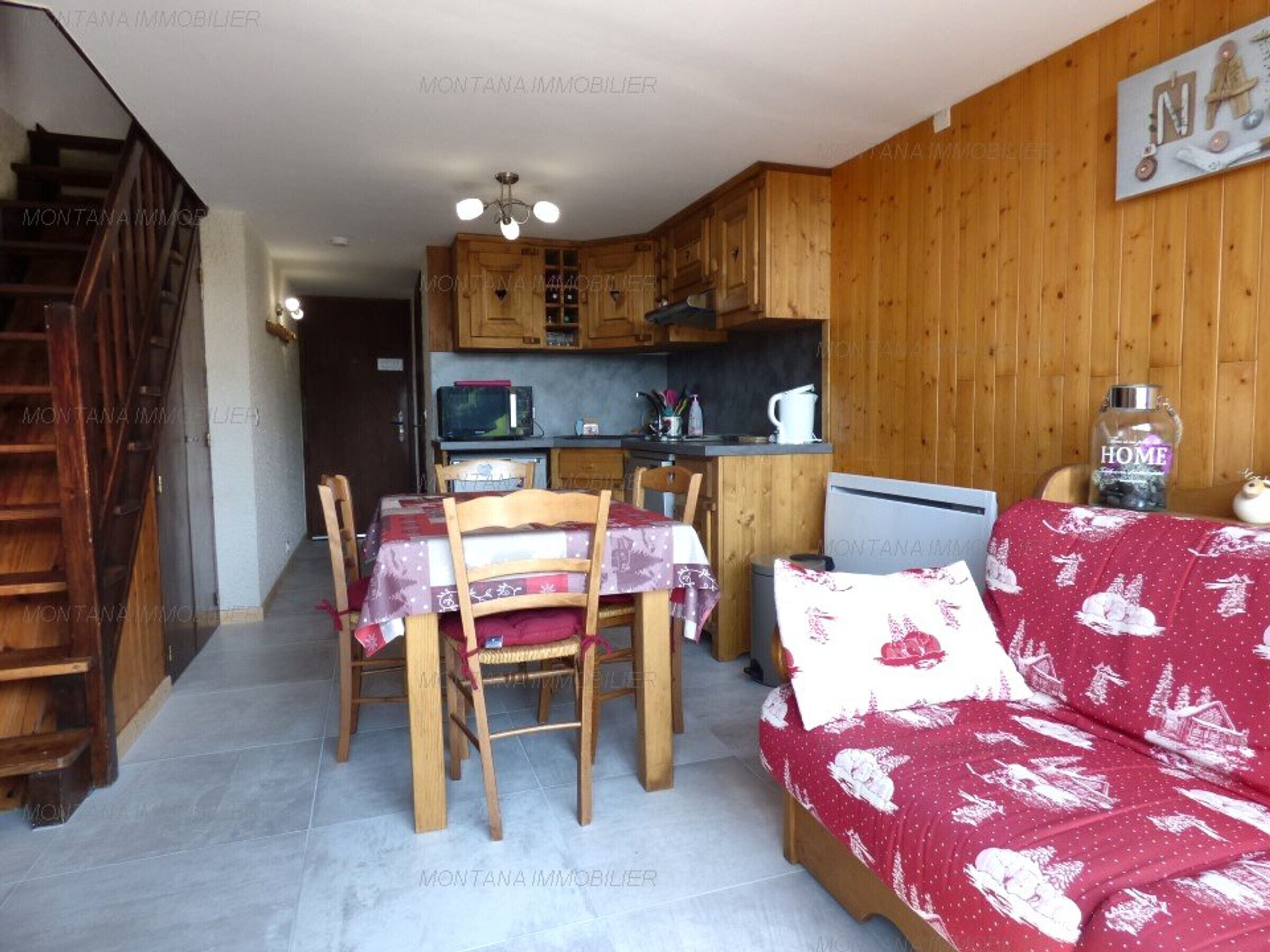 A cozy living room with a wooden staircase, a dining area with a table and chairs, a kitchen with wooden cabinets, and a red sofa with a white pattern.