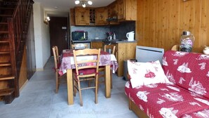A cozy living room with a wooden staircase, a dining area with a table and chairs, a kitchen with wooden cabinets, and a red sofa with a white pattern.
