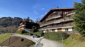 A multi-story building with balconies, a parking area, and a mountainous backdrop.