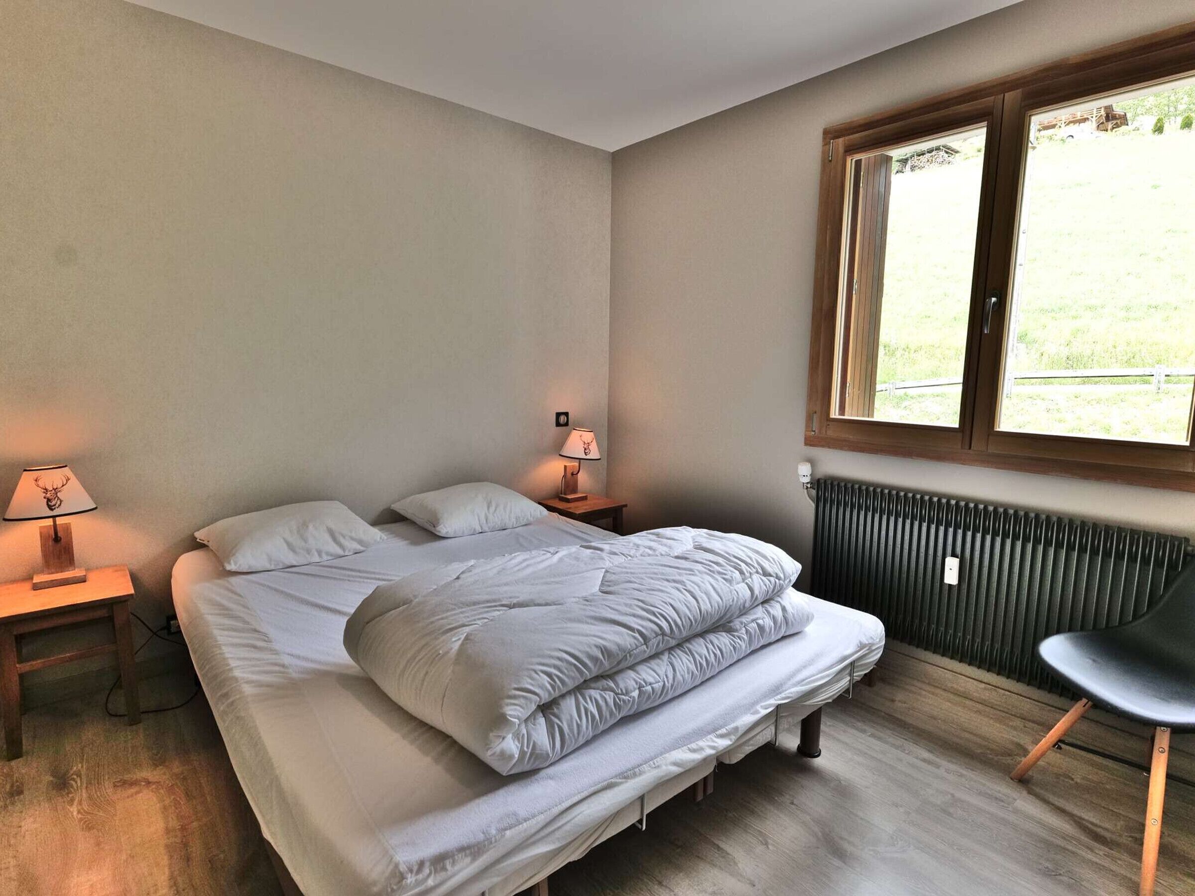 A bedroom with a bed, two bedside lamps, a radiator, a chair, and a window with a view of a grassy area.