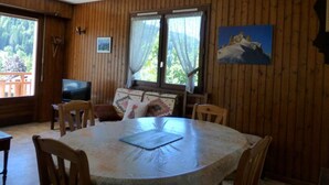 Et rum med træpaneler, et rundt bord, stole, et fjernsyn og et vindue med gardiner.