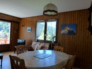 Ein Zimmer mit Holzwänden, einem runden Tisch, Stühlen, einem Fernseher und einem Fenster mit Vorhängen.