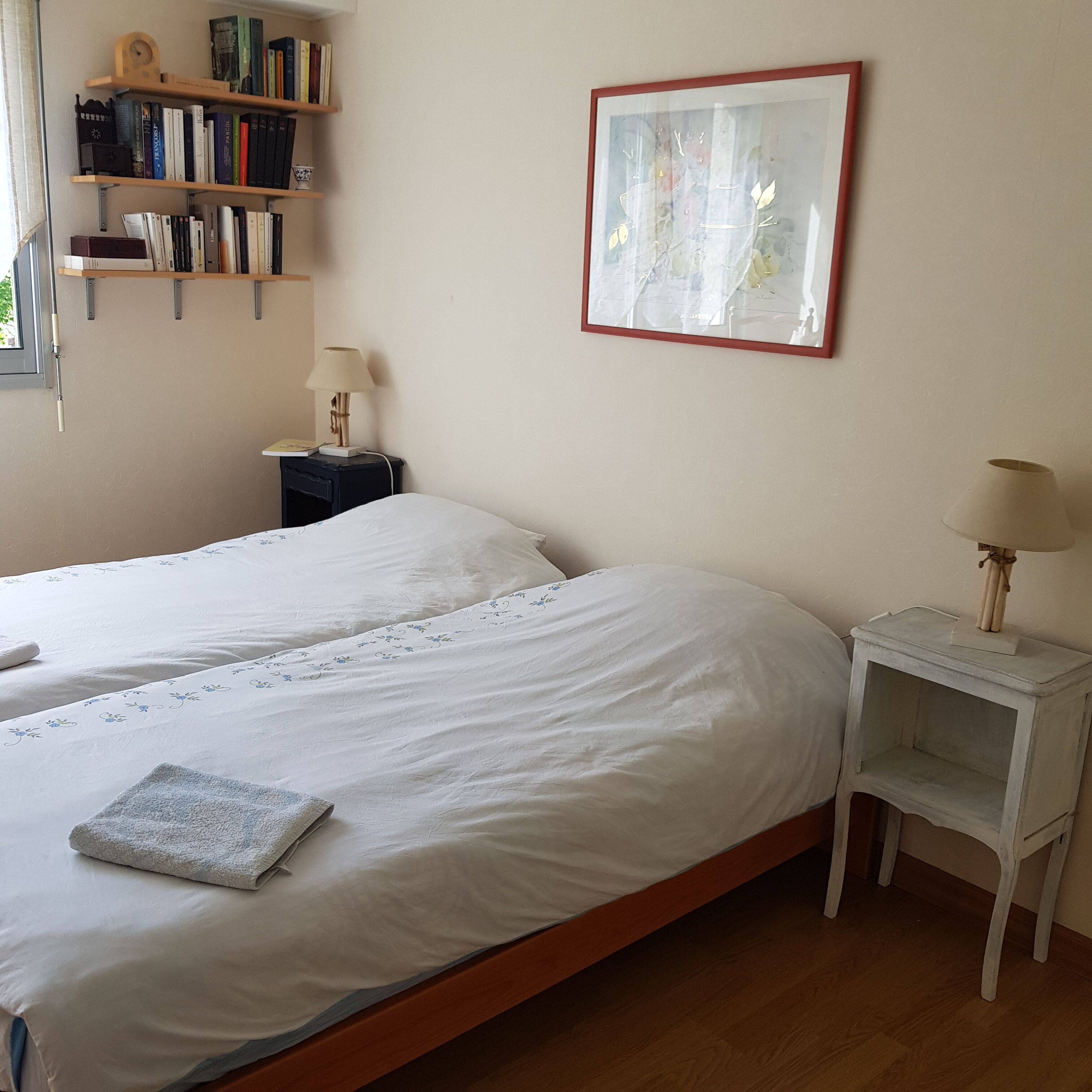 Ein ordentlich gemachtes Bett mit weißen Leinentüchern und einem Blumenmuster, ein Nachttisch mit einer Lampe, ein Bücherregal mit verschiedenen Büchern und ein gerahmtes Bild an der Wand.