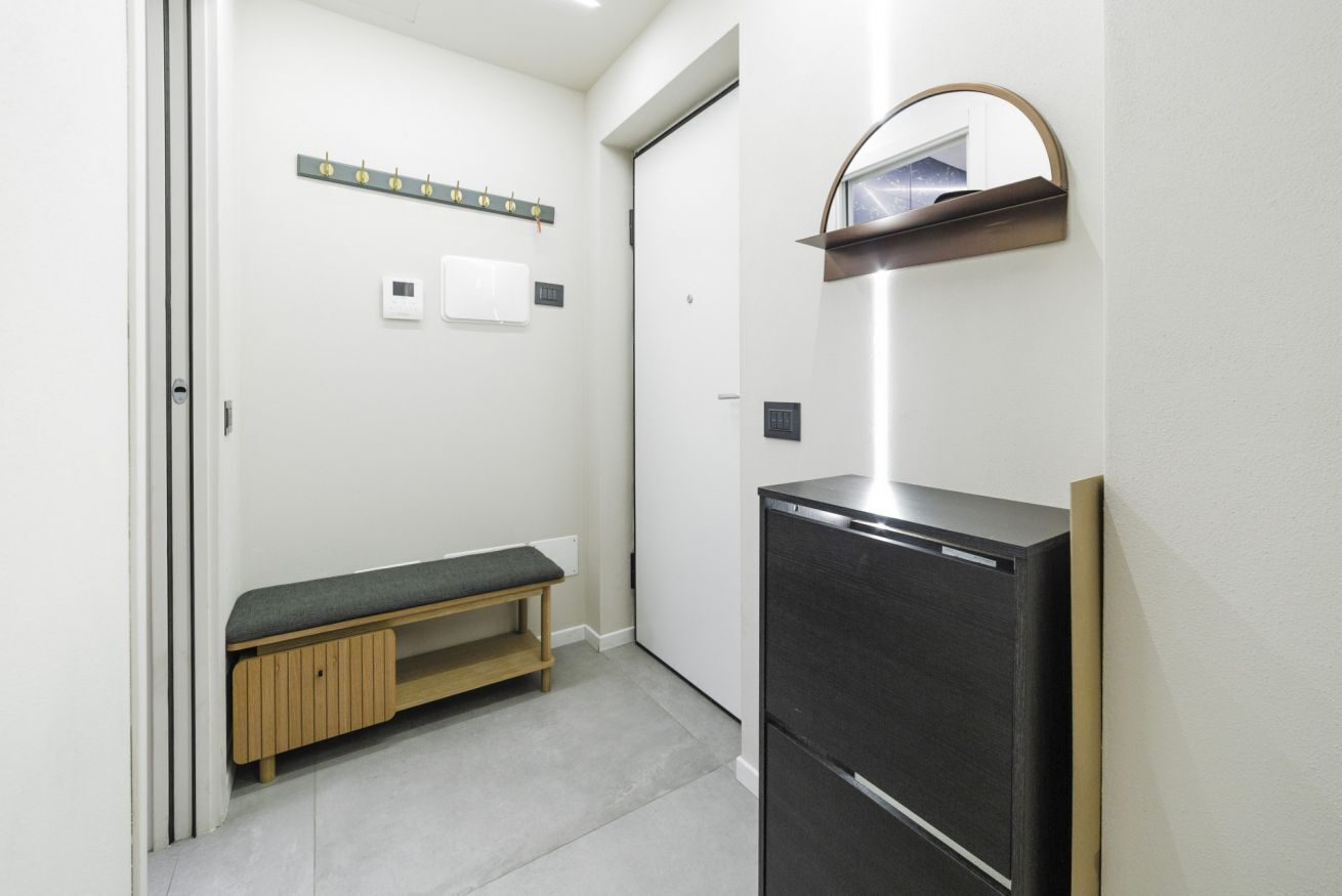 A compact room with a bench, a black refrigerator, a wall-mounted shelf, and a door with a handle.