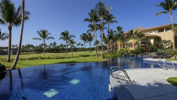 A swimming pool with a clear blue water, surrounded by palm trees and a building with a tiled roof. - Corner Villa with Golf Course Views | Steps from Waikoloa Shops & Beaches (Waikoloa Village)