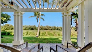 A covered outdoor seating area with wicker chairs, a view of a golf course, and a white pergola structure. - Corner Villa with Golf Course Views | Steps from Waikoloa Shops & Beaches (Waikoloa Village)