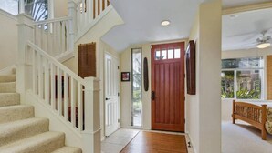 A staircase with white railings leading to a landing, a wooden door, and a bench in a well-lit room with a view of greenery outside. - Corner Villa with Golf Course Views | Steps from Waikoloa Shops & Beaches (Waikoloa Village)