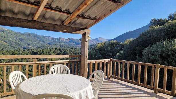 Eine überdachte Terrasse im Freien mit Tisch und Stühlen, mit Blick auf eine gebirgige Landschaft.