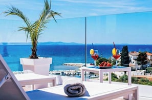 Un balcon offrant une vue dégagée sur la mer, une chaise blanche, une petite table avec un verre, et une serviette enroulée.