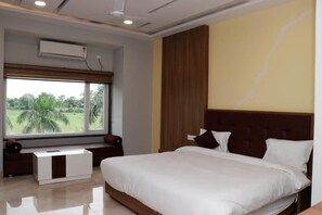 A hotel room with a large bed, a bedside table, a nightstand, a window with a view of greenery, and an air conditioning unit. - Hotel Regal (Jabalpur)