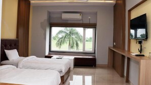 A hotel room with a bed, a desk, a TV, and a window with a view of palm trees. - Hotel Regal (Jabalpur)