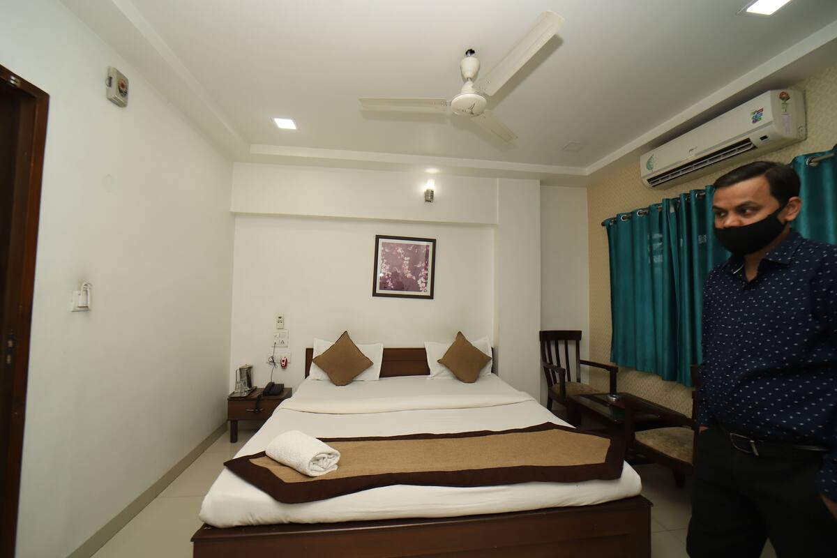 A hotel room with a bed, a ceiling fan, an air conditioning unit, a framed picture on the wall, and teal curtains.