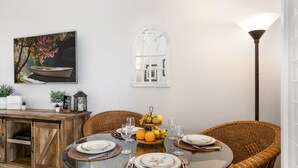 Un coin repas avec une table ronde en verre, dressée pour quatre personnes, entourée de chaises en osier. Un buffet en bois avec une porte coulissante se trouve à gauche, et un cadre photo est accroché au mur au-dessus de la table.