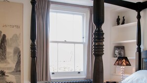 Une chambre à coucher avec un lit à baldaquin, un radiateur, une fenêtre avec des rideaux, une table de chevet avec une lampe et un mur orné d’un tableau encadré.
