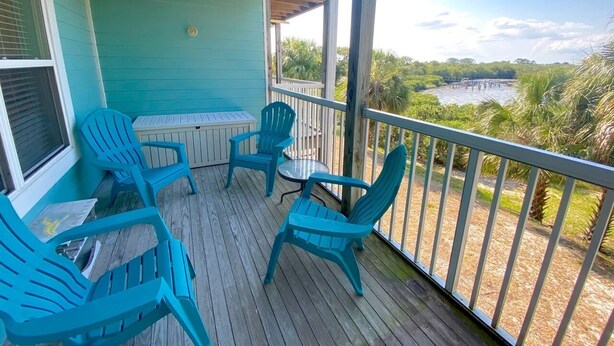Una terraza techada con sillas azules y una mesa, con vista a un cuerpo de agua y follaje.