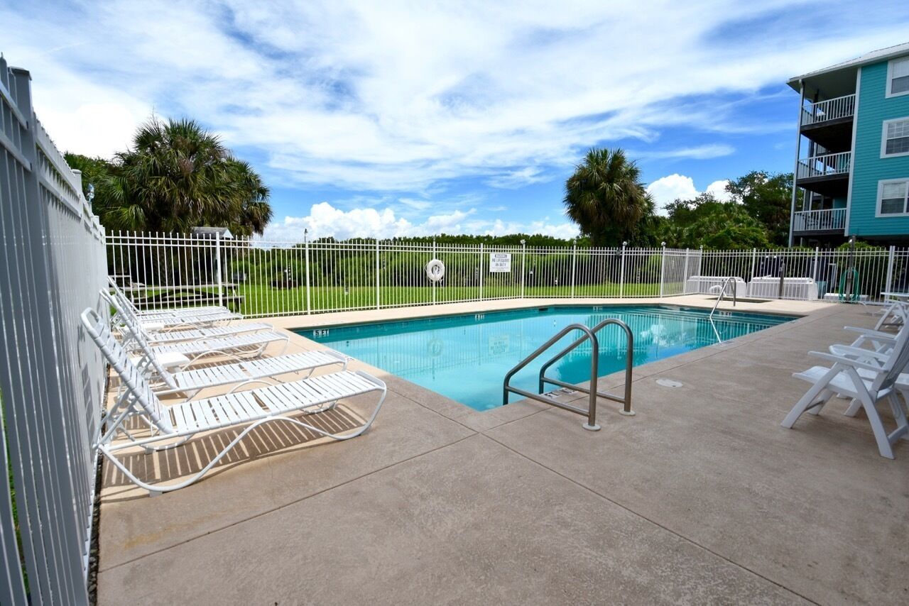 Uma piscina com cercado branco, cadeiras de descanso e um edifício ao fundo.