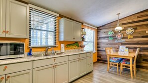 A kitchen with light-colored cabinets, a microwave, and a sink. There is a wooden wall with a clock and a sign that says "here." - Hawthorne - Fisherman's Delight! Plenty of Parking (Cedar Key)