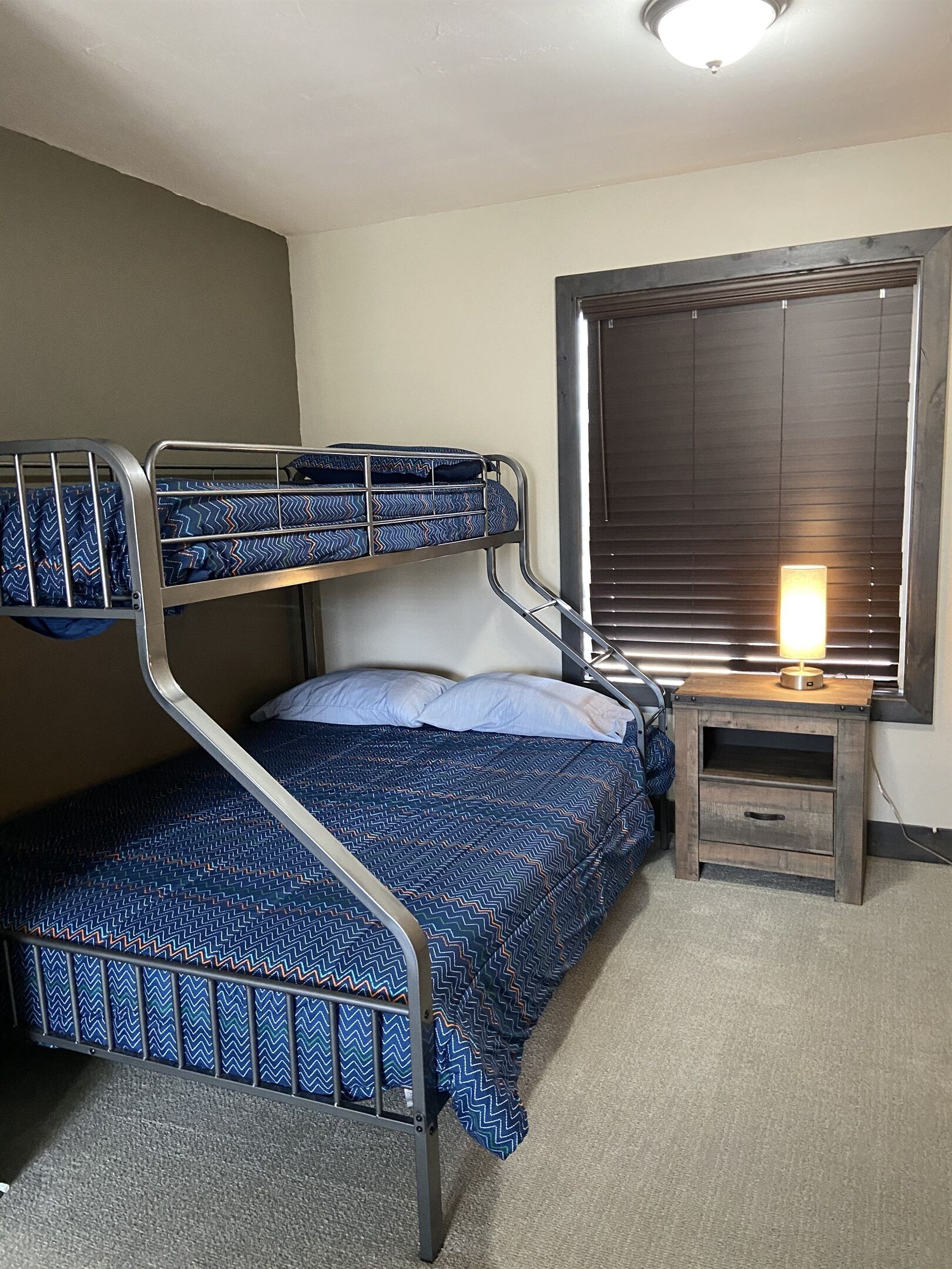 Una cama litera con colchón azul estampado, una mesita de noche con lámpara y una ventana con persianas.
